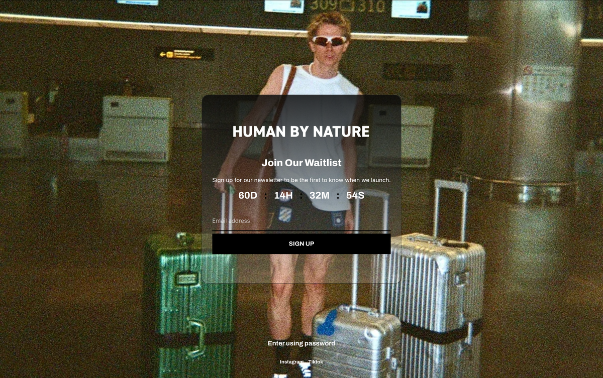 A person standing in an airport terminal with three hard-shell suitcases, wearing sunglasses and a white tank top. A large semi-transparent overlay reads 'HUMAN BY NATURE' with a signup call-to-action.